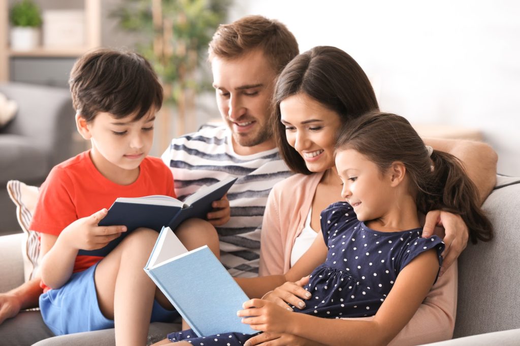 Father, mother, and their two children all sit together while reading books to pass the time at home.